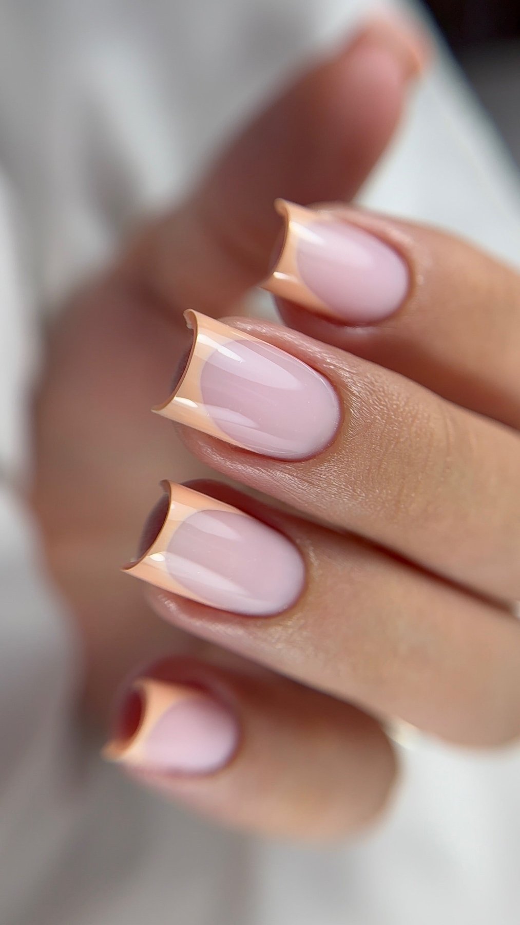 Gros plan macro d'une main avec une manucure de style French manucure bicolore, avec des bases rose poudré et des pointes beiges/abricot, façonnées en un design géométrique pointu et sculpté sur des ongles de forme carrée. Le fond est flou, mettant l'accent sur les détails de l'ongle.