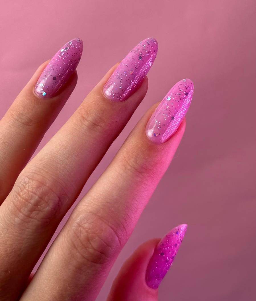 Pose du Gel Polish Xoxo rose parme pailleté sur ongles en amande longs, fond rose