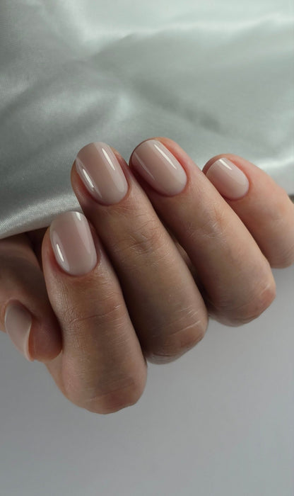 Main gainée au gel Easy gel Toffee devant un satin blanc, ongles courts beige nude
